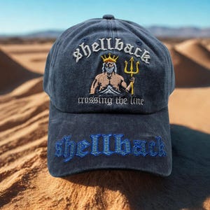 Peut inclure: Casquette de baseball bleu marine avec le mot "shellback" brodé en blanc et bleu. La casquette présente une image brodée d'une figure musclée avec une couronne et un trident, avec le texte "crossing the line".