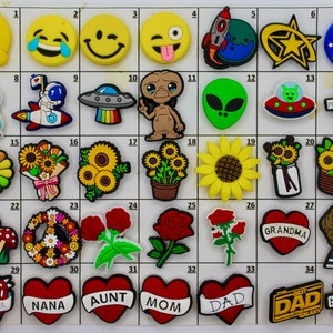 May include: A collection of colorful shoe charms featuring various designs. Includes emojis, space-themed elements like rockets and aliens, floral motifs, and heart-shaped charms with text like "Grandma" and "Dad".