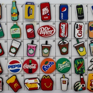 May include: A collection of 35 colorful shoe charms featuring popular beverage brands, including Coca-Cola, Sprite, Dr Pepper, Pepsi, 7 Up, and Monster Energy. The charms are arranged in a grid on a white background.