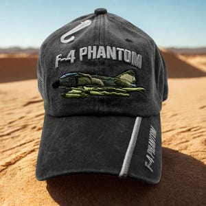 May include: A black baseball cap with the text "F-4 PHANTOM" embroidered in white. The cap features an embroidered image of a military aircraft in green and brown. A white stripe with the text "F-4 PHANTOM" runs down the side.