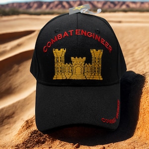 May include: Black baseball cap with "COMBAT ENGINEER" embroidered in red and a gold castle design. The cap has a curved brim and a tag attached. The cap is sitting on sand.