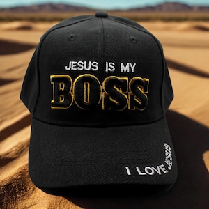 以下が含まれることがあります： 黒い野球帽。白と金色の糸で「JESUS IS MY BOSS」と刺繍されています。側面には「I LOVE JESUS」の文字が刺繍されています。砂漠の背景に配置されています。