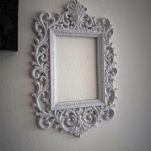 May include: A white, ornate picture frame with detailed scrollwork and a rectangular aperture. The frame is mounted on a light-coloured wall. The frame has a vintage, antique aesthetic.