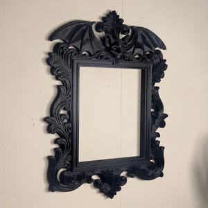 May include: A black, decorative picture frame featuring a bat design. The frame has a rectangular aperture and is adorned with detailed scrollwork and a bat at the top. It is mounted on a light-coloured wall.