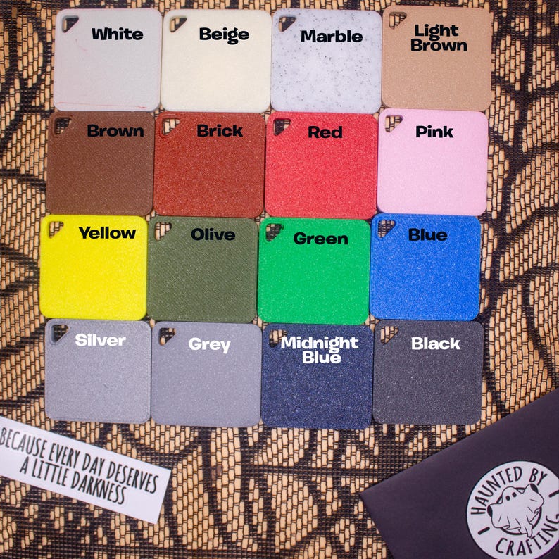 a collection of twelve square tiles, each with a different color, arranged in a grid-like pattern. The tiles are labeled with the names of the colors, such as "White", "Beige", "Marble", "Light Brown", "Pink", "Yellow", "Olive", "Green", "Blue", "Silver", "Grey", "Midnight Blue", and "Black". The tiles are placed on a textured surface, and there is a small black object in the bottom right corner.