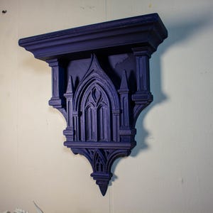 May include: A dark purple, gothic-style wall shelf. The shelf features an arched window design with columns and decorative elements. It is mounted on a white wall, showcasing its intricate details and architectural style. The shelf is a decorative piece.