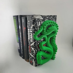 May include: A green tentacle bookend supports a stack of books. The bookend is a 3D-printed design with a detailed, textured surface. The books have various titles and colors, with one featuring the text "LA PORTA OSCURA".