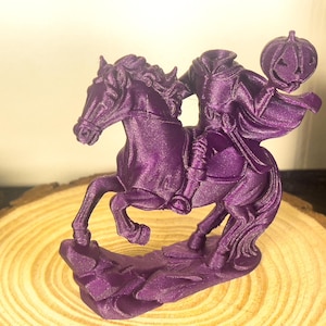 May include: A purple 3D-printed sculpture of the Headless Horseman riding a horse, holding a jack-o'-lantern. The sculpture is on a wooden slice. The horse is in mid-gallop, and the figure is cloaked.