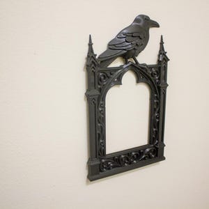 May include: A black decorative wall frame with a raven perched on top. The frame has an arched top and ornate detailing, creating a gothic aesthetic. The raven is also black, with detailed feathering.