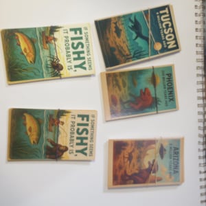 Może przedstawiać: Kolekcja zabytkowych pudełek z zapałkami z kolorowymi ilustracjami. Niektóre przedstawiają ryby i tekst "If something seems fishy, it probably is." Inne wyświetlają "Tucson", "Phoenix" i "Arizona" z "million years ago."