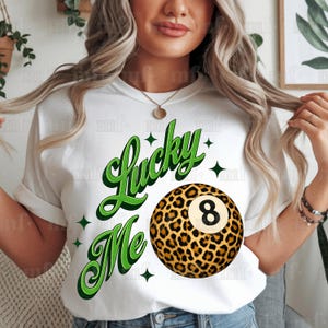 Op de afbeelding: Wit T-shirt met de tekst "Lucky Me" in groen schrift en een achtballenafbeelding met luipaardprint. Het ontwerp bevat groene steraccenten. Een casual kledingstuk voor dagelijks gebruik.