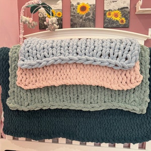 May include: Four chunky knit blankets stacked on a white crib. The blankets are in shades of blue, pink, green, and dark blue.