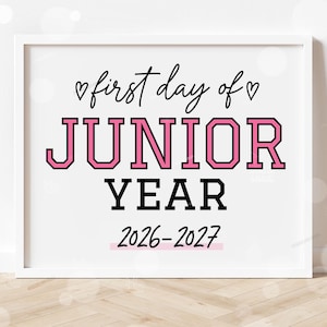 May include: A white framed sign featuring the black text first day of with two small hearts, the word JUNIOR in large pink block letters with black outlines, the word YEAR in black, and the school year 2026-2027 underlined in light pink.
