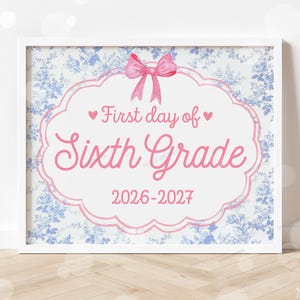 May include: A framed sign featuring a blue and white floral background with a pink scalloped border and a pink bow at the top. The text reads First day of Sixth Grade 2026-2027 in pink script and block lettering with small pink hearts on the sides.