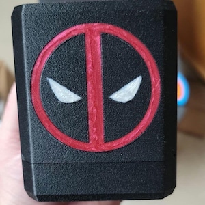 Könnte beinhalten: Eine schwarze Box mit einem roten Deadpool-Logo. Das Logo ist ein Kreis mit rotem Rand und schwarzem Inneren. Die Augen sind weiß.