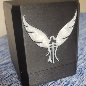 Op de afbeelding: Zwarte kubusvormige doos met een wit gevleugeld engelontwerp aan de voorkant. De engel heeft een zwaard in zijn hand.