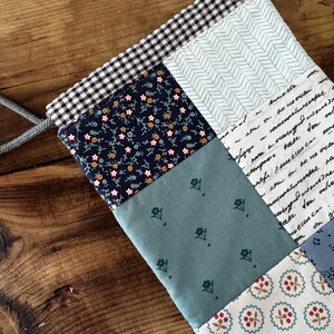 Può includere: Una borsa con coulisse caratterizzata da un design patchwork con vari motivi in tessuto, tra cui quadretti vichy bianchi e neri, fiori blu navy, spina di pesce azzurra, testo scritto in bianco, verde salvia con piccoli fiori e bianco con motivi a bacche rosse.