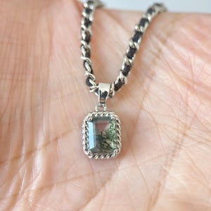 May include: A silver pendant necklace featuring a rectangular, green-hued gemstone with a unique, natural pattern. The pendant is framed with a twisted rope design and hangs from a black and silver chain.
