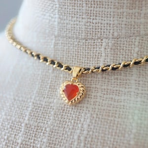 May include: Gold-coloured necklace with a heart-shaped pendant. The pendant has a red gemstone set in a decorative gold frame. The necklace chain combines gold links and black cord.