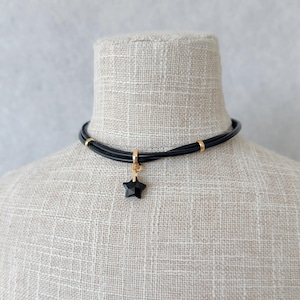 May include: Black choker necklace with a gold-coloured star pendant. The necklace features a multi-strand black cord with gold-coloured accents. The star pendant is a faceted, dark-coloured gemstone.