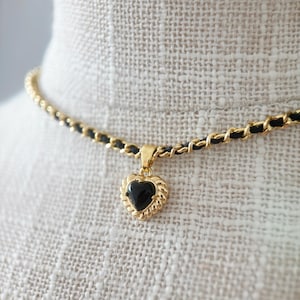 Può includere: Una collana color oro con un ciondolo a forma di cuore nero. Il cuore è incorniciato da un bordo dorato e pende da una catena con dettagli neri. La collana è esposta su una superficie di colore neutro.