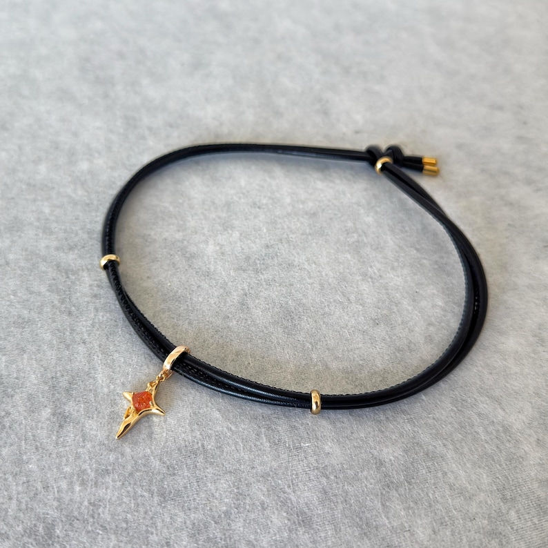 May include: Black leather bracelet with a gold-colored star charm. The star has an orange center and is suspended from a gold-colored clasp. The bracelet has gold-colored accents and a sliding knot closure.