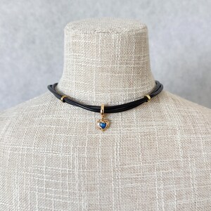 May include: A black choker necklace with a gold-colored heart pendant. The pendant features a blue gemstone in a heart-shaped frame. The necklace is displayed on a mannequin with a neutral background.