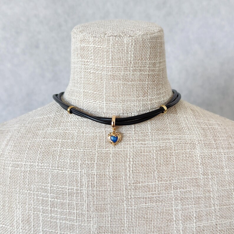 May include: A black choker necklace with a gold-colored heart pendant. The pendant features a blue gemstone in a heart-shaped frame. The necklace is displayed on a mannequin with a neutral background.