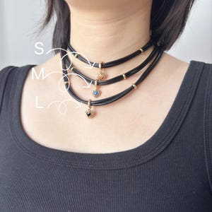 May include: Three black choker necklaces with heart-shaped pendants in gold settings. Each pendant features a different colored stone: peach, blue, and black. The necklaces are labeled S, M, and L.