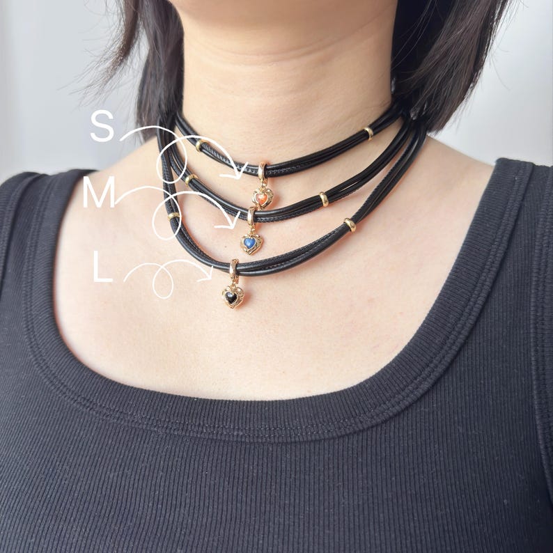May include: Three black choker necklaces with heart-shaped pendants in gold settings. Each pendant features a different colored stone: peach, blue, and black. The necklaces are labeled S, M, and L.