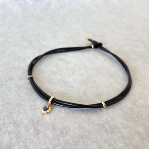 May include: Black cord bracelet with a gold-colored heart-shaped charm. The charm has a blue center and a textured border. The bracelet also features gold-colored accents.