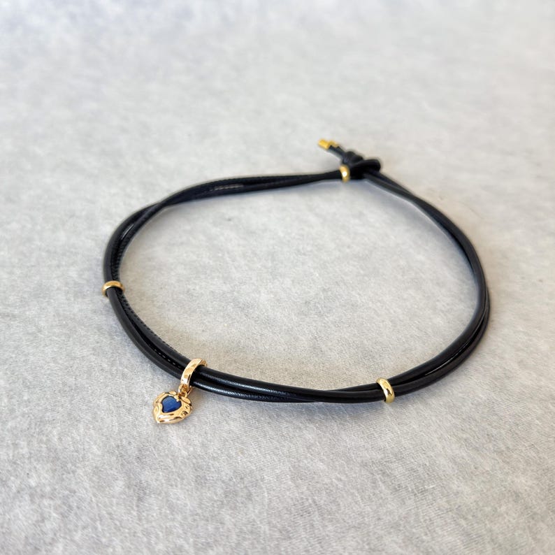 May include: Black cord bracelet with a gold-colored heart-shaped charm. The charm has a blue center and a textured border. The bracelet also features gold-colored accents.