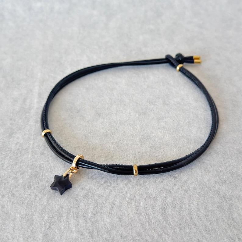 May include: Black bracelet with a star charm. The bracelet has a black cord design with gold-coloured accents and a small, dark star pendant. The bracelet is knotted at the closure.