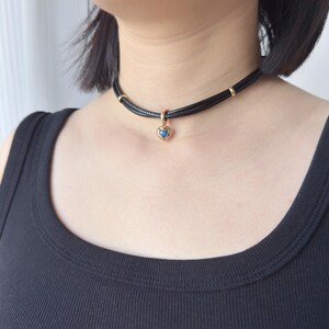 May include: Black choker necklace with a gold heart pendant. The pendant features a blue gemstone center. The necklace is made of black leather and has gold accents. A stylish piece of jewelry.