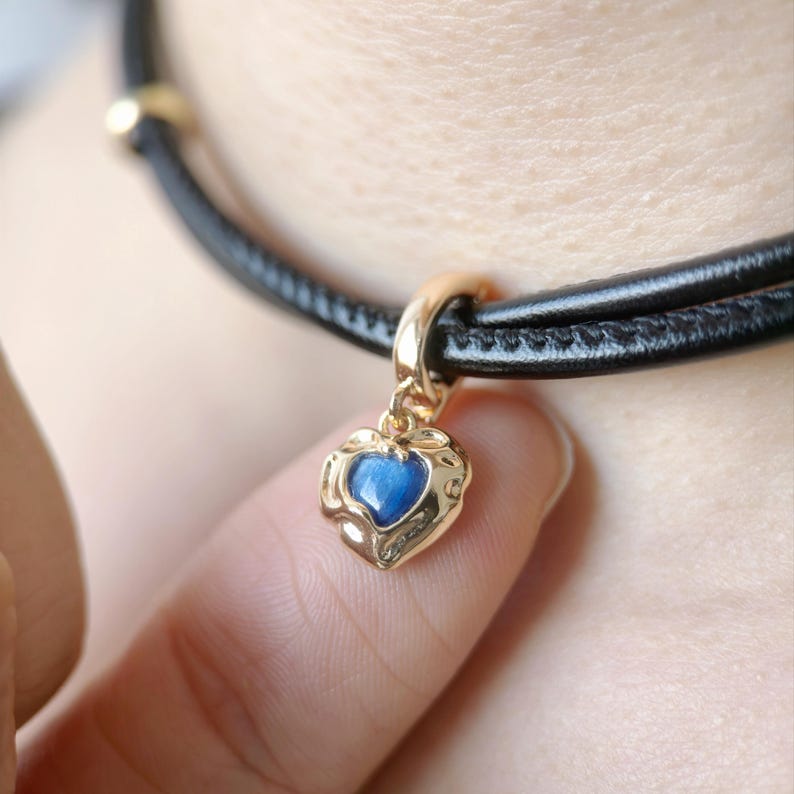 May include: A gold-colored heart-shaped pendant with a blue center stone hangs from a black leather cord necklace. The pendant is detailed with a floral design around the heart. The necklace is worn around the neck.
