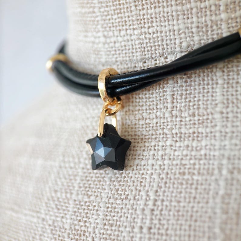May include: Black star pendant necklace. The necklace features a black star-shaped pendant with a faceted design, suspended from a gold-coloured clasp. The necklace has a black leather cord with gold-coloured accents.