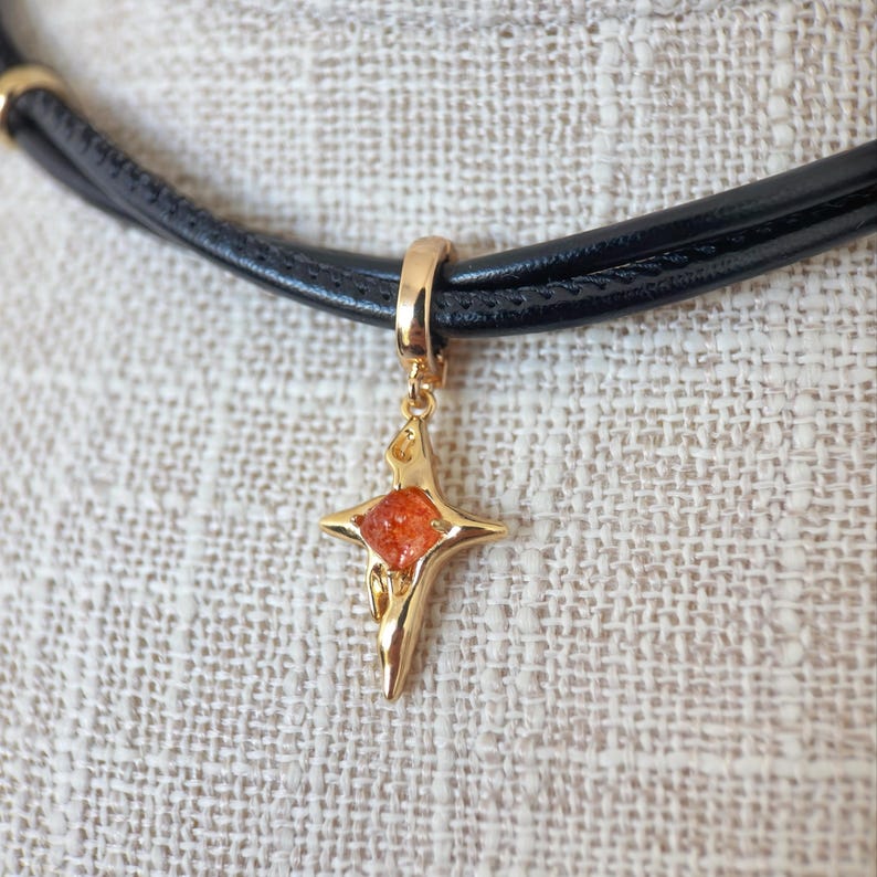 May include: A gold-colored pendant in the shape of a star with an orange center, suspended from a black leather choker necklace. The necklace has a triple-strand design and a gold-colored clasp. The pendant is approximately 1 inch in length.