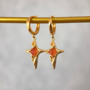 May include: A pair of gold-colored hoop earrings. Each earring features a starburst pendant with an orange, gem-like center. The earrings are displayed on a gold bar against a soft, neutral background. The earrings are designed to be worn.