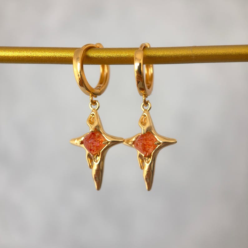 May include: A pair of gold-colored hoop earrings. Each earring features a starburst pendant with an orange, gem-like center. The earrings are displayed on a gold bar against a soft, neutral background. The earrings are designed to be worn.