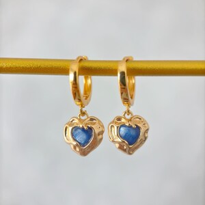 May include: A pair of gold-colored hoop earrings with heart-shaped pendants. Each pendant features a textured gold frame surrounding a blue center. The earrings are displayed on a gold bar against a soft, neutral background.