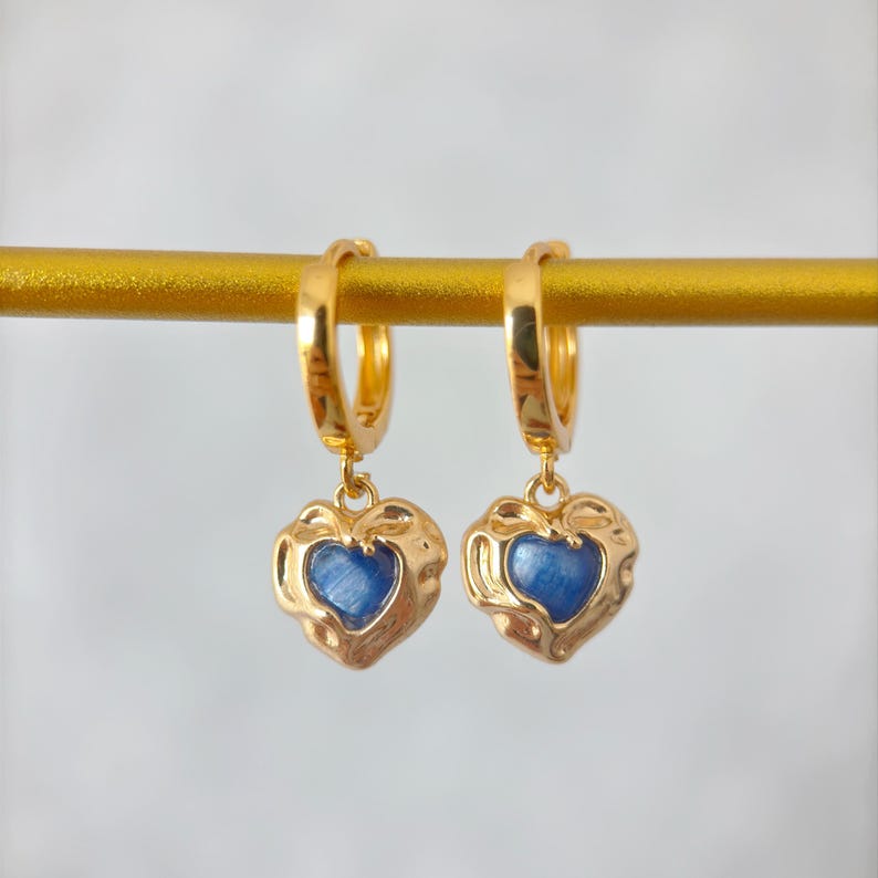 May include: A pair of gold-colored hoop earrings with heart-shaped pendants. Each pendant features a textured gold frame surrounding a blue center. The earrings are displayed on a gold bar against a soft, neutral background.