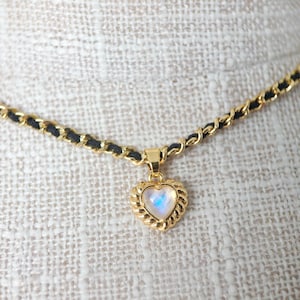 May include: A gold-toned necklace with a black woven chain. The pendant is a heart-shaped charm with a gold rope-like border and a pearlescent center. The necklace is displayed on a textured, off-white surface.