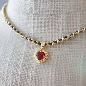 Può includere: Una collana dorata con catena nera e pendente a forma di cuore. Il pendente presenta una pietra rossa incastonata in una cornice dorata decorativa. La collana è esposta su uno sfondo di tessuto neutro.