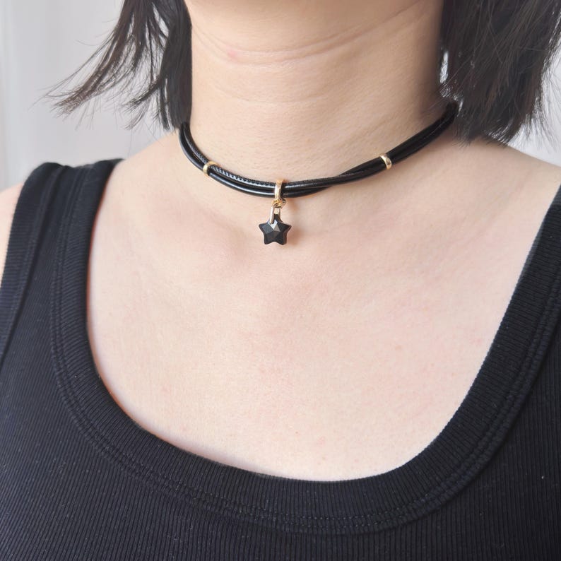 May include: Black choker necklace featuring a star-shaped pendant. The necklace has a thin, black cord with gold-coloured rings and a gold-coloured clasp. The star pendant is a dark, faceted stone.