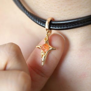 May include: A gold-colored pendant in the shape of a star with an orange gemstone center, suspended from a black leather choker necklace. The pendant is held by a finger, showcasing the jewelry's design and color.