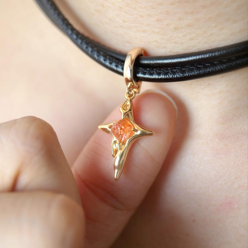 May include: A gold-colored pendant in the shape of a star with an orange gemstone center, suspended from a black leather choker necklace. The pendant is held by a finger, showcasing the jewelry's design and color.