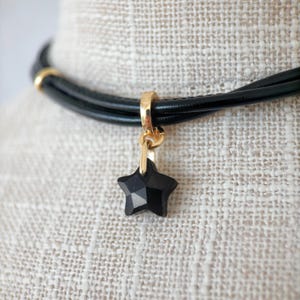 May include: Black leather choker necklace with a gold-coloured clasp and a faceted black star pendant. The necklace has a simple, elegant design, suitable for everyday wear or special occasions.