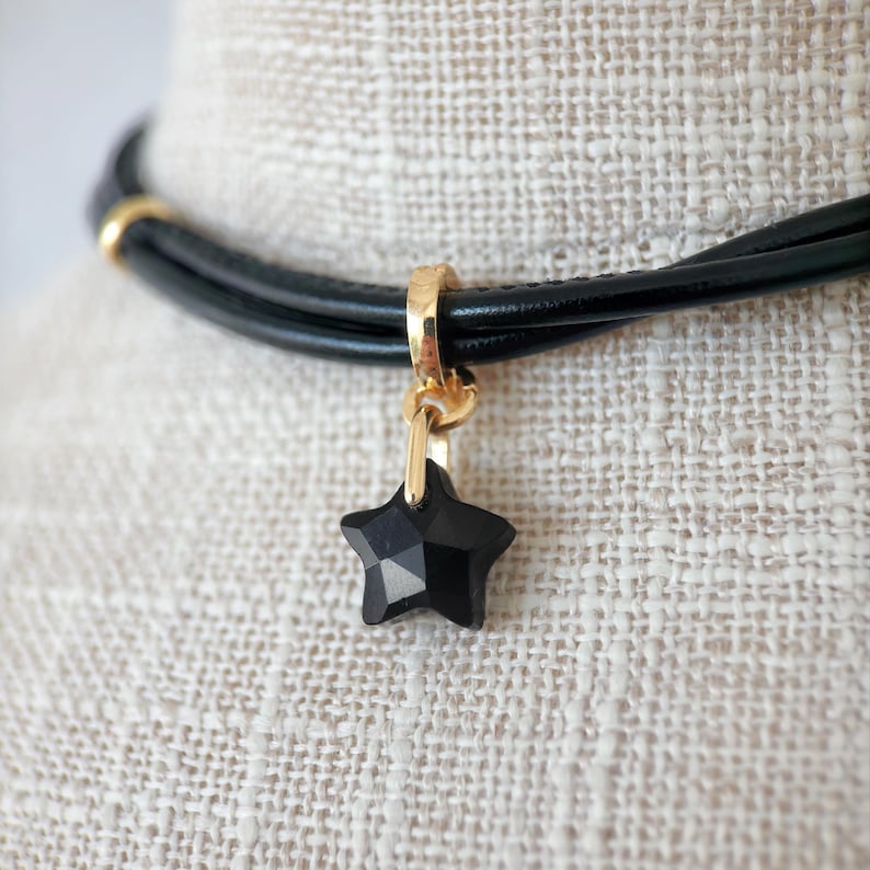 May include: Black leather choker necklace with a gold-coloured clasp and a faceted black star pendant. The necklace has a simple, elegant design, suitable for everyday wear or special occasions.