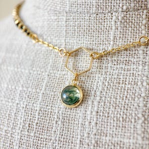 Op de afbeelding: Een goudkleurige ketting met een ronde, groene hanger. De hanger is gezet in een gouden bezel en hangt aan een gouden zeshoekige vorm. De ketting is ook goudkleurig en heeft een delicaat ontwerp. De achtergrond is een neutrale stof.