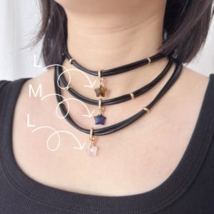 May include: Three black choker necklaces with star-shaped pendants in brown, blue, and pink. Each necklace features gold-coloured accents and a clasp. The necklaces are layered and worn around the neck.